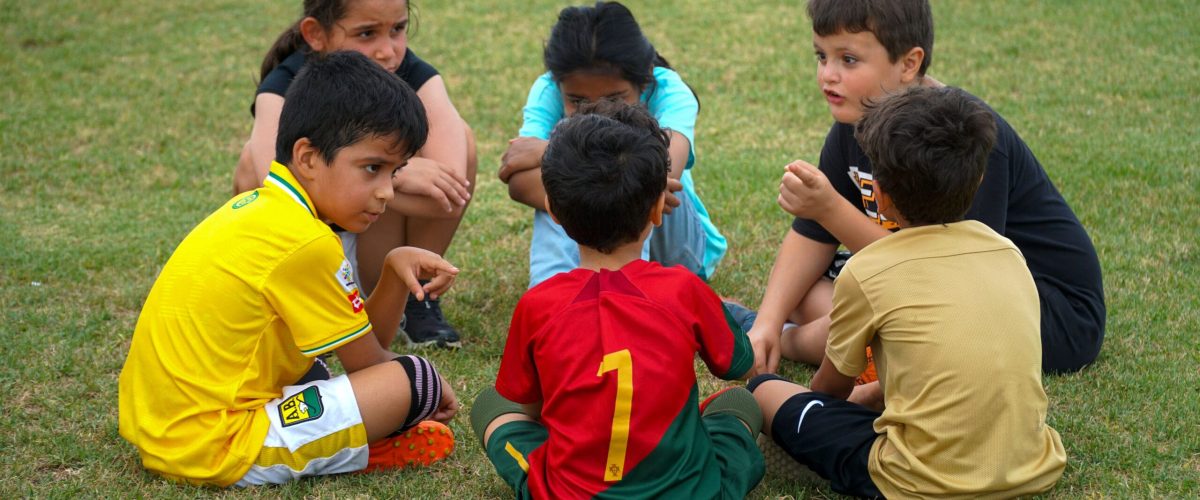 Children sit in a circle on a soccer field during a Child Safety Soccer Tournament.