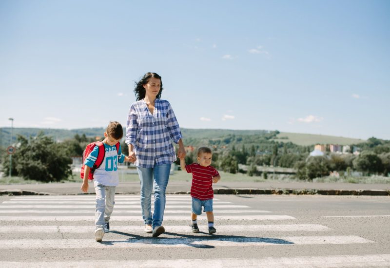 Mother Crossing the Street with Her Children A woman holding hands with two children as they cross a pedestrian crosswalk on a sunny day, with a rural landscape in the background.