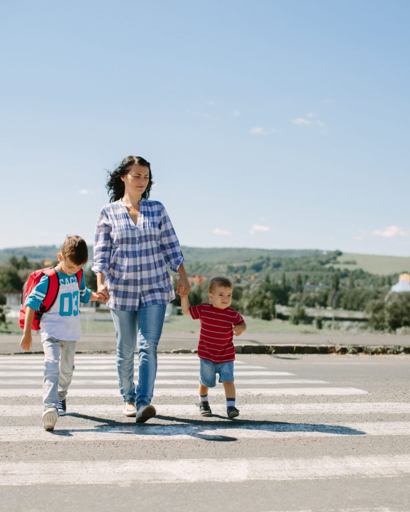 Mother Crossing the Street with Her Children A woman holding hands with two children as they cross a pedestrian crosswalk on a sunny day, with a rural landscape in the background.