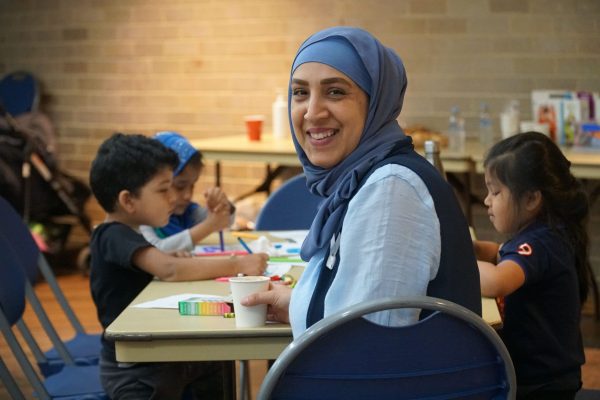 A smiling woman in a hijab sits at a table with children engaged in arts and crafts, reflecting community values of learning and inclusion.