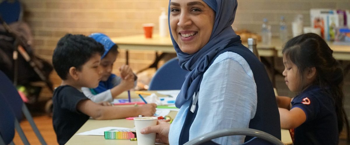Our Values: Community, Learning & Inclusion A smiling woman in a hijab sits at a table with children engaged in arts and crafts, reflecting community values of learning and inclusion.